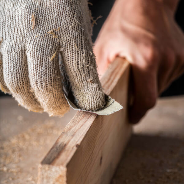 Gros plan d'une main gantée ponçant le bord d'une planche en bois avec du papier de verre.