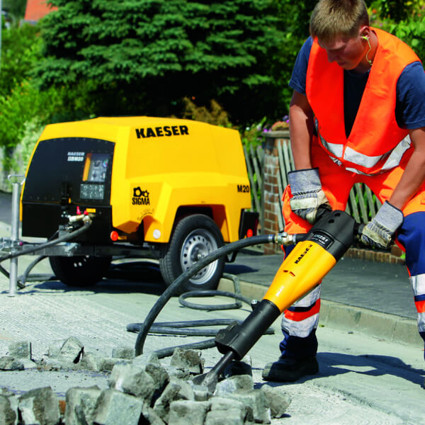 Un ouvrier en gilet de sécurité utilisant un marteau-piqueur raccordé à un compresseur Kaeser.