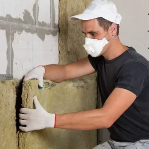 Un homme portant un masque et des gants installe de l'isolation dans un espace de construction.