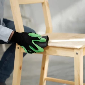 Une personne porte un gant vert et nettoie une chaise en bois avec un chiffon.